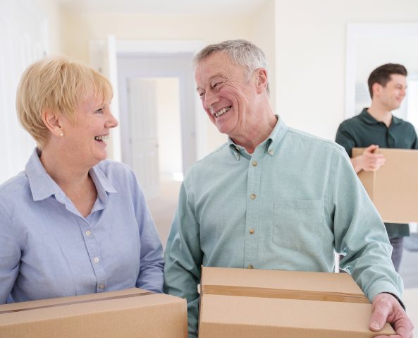 Senior Couple Downsizing In Retirement Carrying Boxes Into New Home On Moving Day With Removal Man Helping